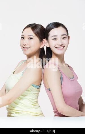 Two young Asian women with their backs to each other, facing the camera, smiling, wearing tank tops, on a white background. Stock Photo