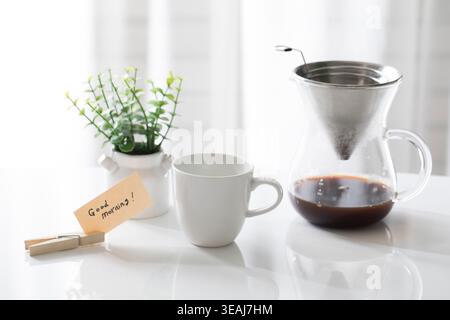A coffee maker sits on a white table, pouring coffee into a cup, in a bright and airy room. Stock Photo
