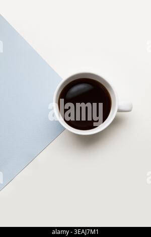 A white coffee cup filled with dark coffee sits on a gray and white paper surface. Stock Photo