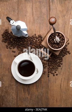 A coffee cup, coffee beans, and a Moka pot sit on a wooden table, surrounded by a rich, dark coffee. Stock Photo