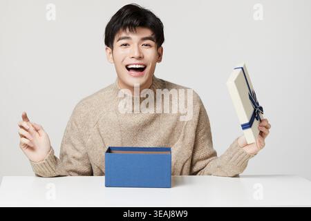 A young Asian man excitedly opens a gift box on a white table. Stock Photo