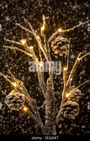 A snow-covered Christmas tree branches with pinecones and fairy lights lit up in the snowfall. Stock Photo