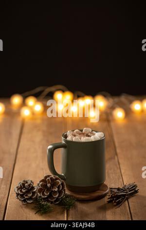 A steaming mug of hot chocolate sits on a wooden table, surrounded by festive pinecones and twinkling fairy lights. Stock Photo