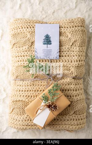 A cozy, hand-knitted cream-colored blanket is gift-wrapped with a small present and a card on a soft, white, textured background. Stock Photo