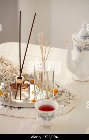 A glass table setting with a teapot, cups, and reed diffuser bottles, arranged on a white tablecloth. Stock Photo