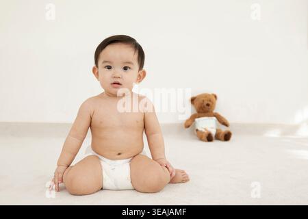 A baby sits on a white floor with a teddy bear in the background. Stock Photo