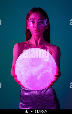 A woman in futuristic glasses holds a glowing moon orb in a dark, neon-lit environment. Stock Photo