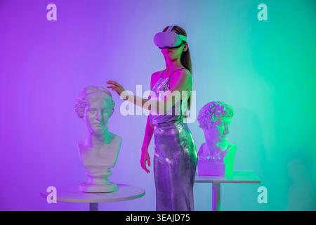 A woman in a futuristic outfit, wearing a VR headset, interacts with two classical busts in a dimly lit, neon-lit room. Stock Photo