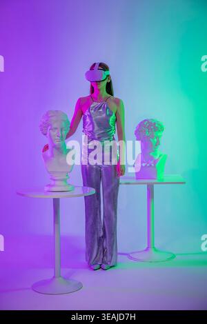 A woman in a metallic jumpsuit stands among ancient busts, wearing a VR headset and exploring virtual art. Stock Photo