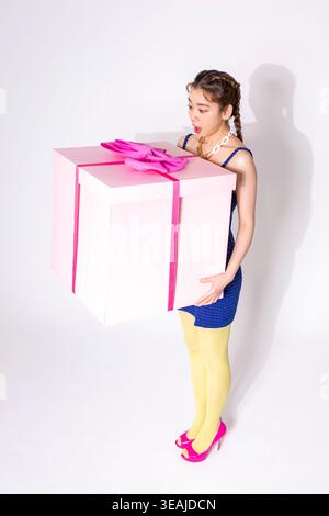 A young woman in a blue dress holds a large pink gift box, her face filled with surprise. Stock Photo