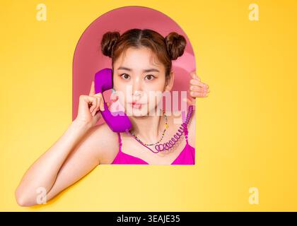 A young woman in a bright pink dress holds a vintage-style purple phone to her ear, smiling in a cheerful, 80s-inspired setting. Stock Photo