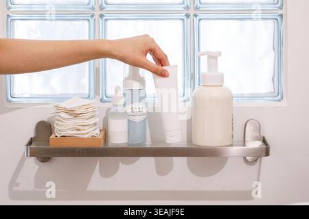A hand reaches for a tube of soap on a bathroom shelf, with a stack of clean towels and a glass window in the background. Stock Photo