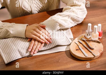 A woman's hands are gently placed on a table, ready for a manicure with various nail care tools and a selection of nail paints nearby. Stock Photo