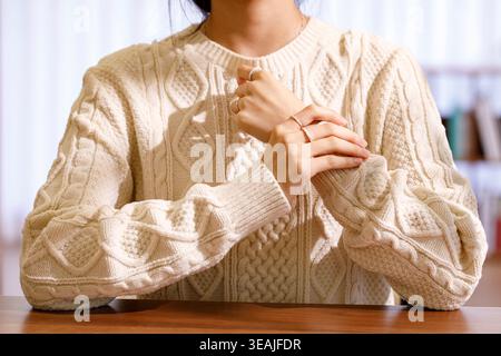 A woman wearing a cream-colored cable-knit sweater sits at a wooden table, her hands clasped in front of her, fingers intertwined. Stock Photo