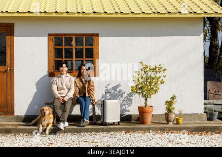 A young couple and their dog sit on a bench outside a cozy cottage, enjoying the warm sunshine. Stock Photo