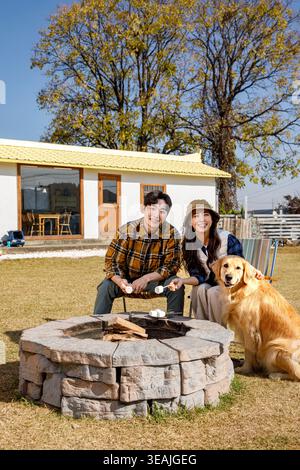 A man and a woman sit by a campfire, roasting marshmallows with their golden retriee, in a sunny backyard. Stock Photo