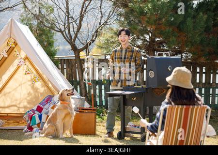 A man and a woman are camping with their dog in a sunny backyard, roasting marshmallows over a grill. Stock Photo