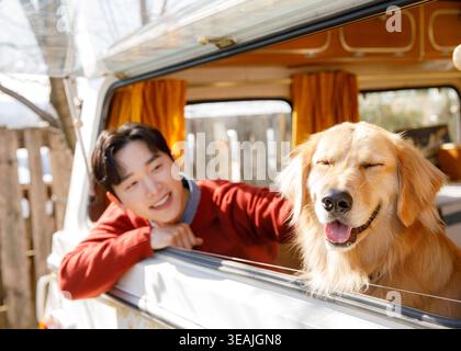 A young man and a happy dog are standing in the back of a vintage camper van, smiling and looking out at the scenery. Stock Photo