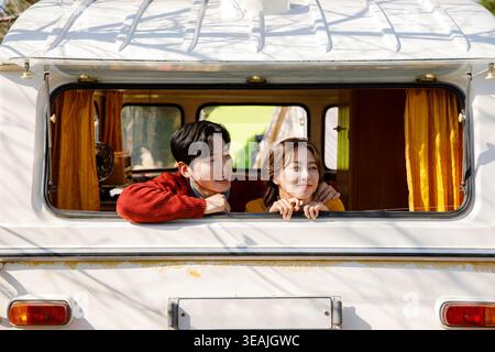 A young couple is smiling and leaning out of a white camper van window, looking out at a sunny landscape. Stock Photo