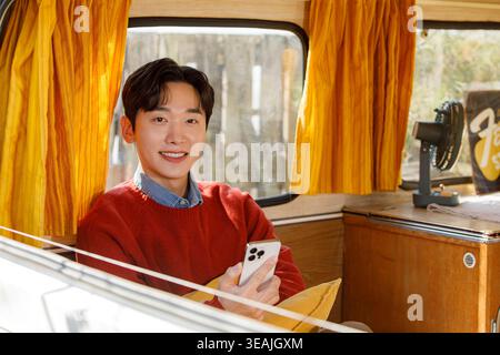 A young man sits in a vintage bus, smiling while holding his phone and looking out the window. Stock Photo