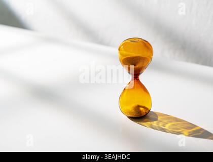 Amber glass hourglass measuring time on a white surface with sunlight and shadows Stock Photo