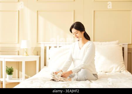 A woman sits on a bed, folding clothes in a cozy bedroom. Stock Photo