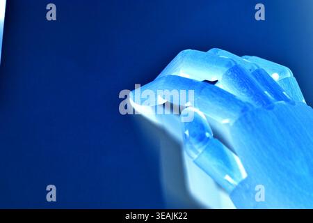 Close-up top view of a single neon blue robotic hand isolated on a blue background. Futuristic minimal concept representing artificial intelligence, r Stock Photo