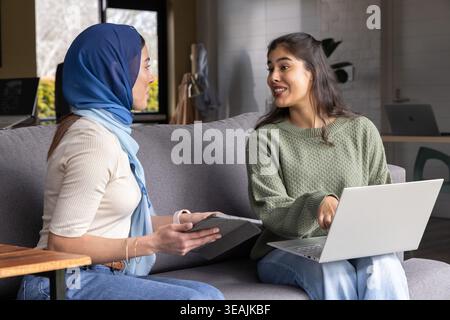 Diverse female colleagues working while pointing at silver laptop on lap and tablet in lounge Stock Photo