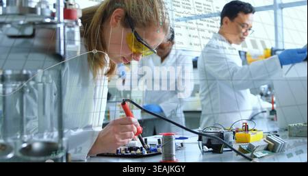 Using soldering iron and spool, researcher leaning over circuit board at bench, wearing lab coat Stock Photo
