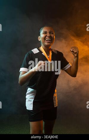 Female athlete celebrating on turf in black-white kit, holding gold medal amid blue-orange smoke Stock Photo