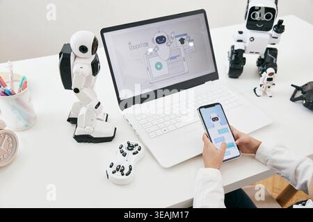 A person sits at a desk, surrounded by various robot models, using a smartphone to control a robot's screen on their laptop. Stock Photo