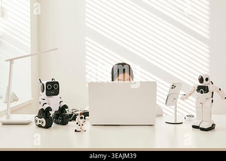 A woman sits at a desk surrounded by robots, working on her laptop. Stock Photo