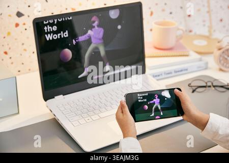 A person uses a smartphone to view a stock image of a laptop screen displaying a 3D animation of a person in a virtual world. Stock Photo