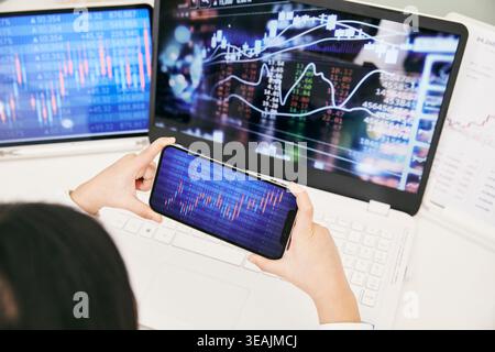 A person is holding a smartphone in front of multiple screens displaying stock market data and charts. Stock Photo
