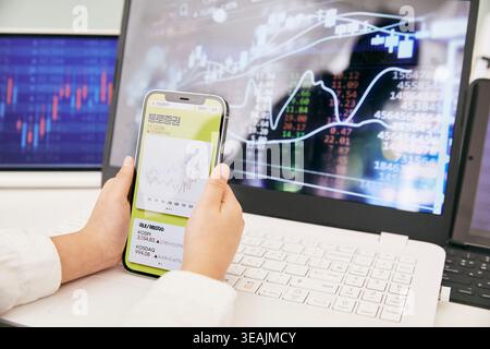 A person's hands are holding a smartphone displaying a stock market app, with a laptop screen and another monitor in the background, showing stock charts and graphs. Stock Photo