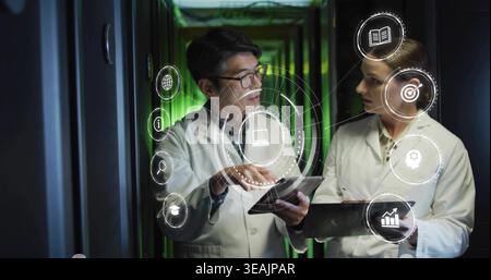 Pointing two technicians in lab coats examining tablets by server racks with green LEDs, AR icons Stock Photo