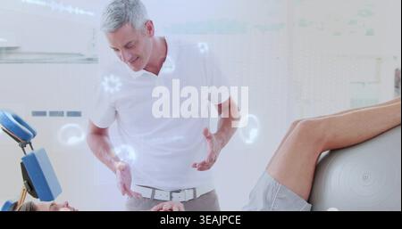 Leaning clinic physio in polo gesturing toward patient on table with gray shorts and ball overlays Stock Photo