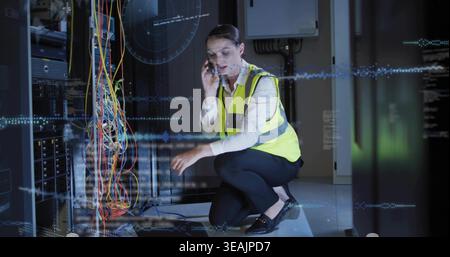 Kneeling technician phoning and checking cables in data center, wearing high-visibility vest Stock Photo