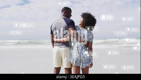 Standing couple embracing on beach, wearing two-tone tee and patterned dress, social icons floating Stock Photo