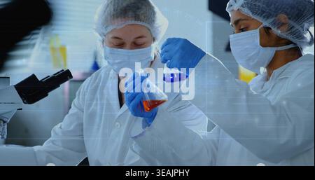 Mixing orange and blue liquids, two techs wearing coats, blue gloves holding flasks at lab bench Stock Photo