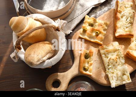 Assorted italian bread with focaccia and olives on wooden board. Top view of fresh bakery products including rolls in basket and sliced flatbread with Stock Photo