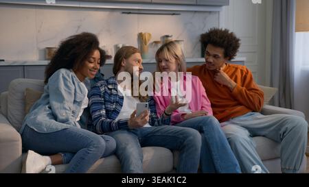 Multiethnic students friends men and women looking at mobile phone at home couch together Caucasian girls African American guys talking chatting news Stock Photo