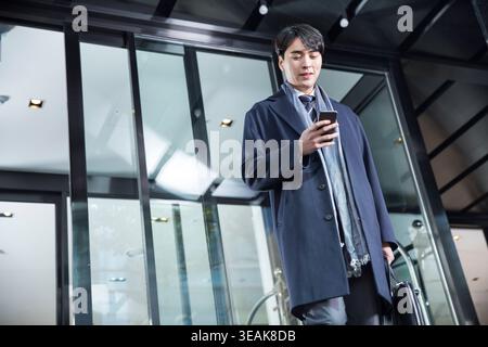 A young businessman stands in a modern office building lobby, checking his phone while holding a briefcase. Stock Photo