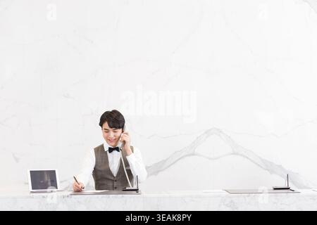 A young male receptionist answers a phone call at a modern white marble reception desk. Stock Photo
