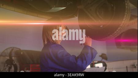 Inspecting auto tech in blue coveralls and mask holding clipboard under raised vehicle in garage. Mechanic, female, ppe, inspection, suspension, wheel Stock Photo