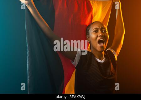 Woman holding large tricolor flag, shouting in studio wearing dark jersey and hoops. Banner, stage, spotlight, teal, amber, haze, dramatic Stock Photo