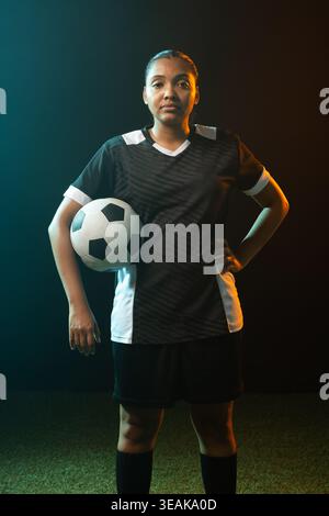 African American woman posing, holding soccer ball on turf in black kit, teal-orange rim light. Athlete, jersey, uniform, kneehigh, portrait, studio, Stock Photo