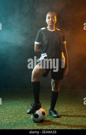 Female soccer player standing on turf wearing black-jersey, foot on ball amid teal-amber smoke. Athlete, studio, portrait, duotone, dramatic, lighting Stock Photo