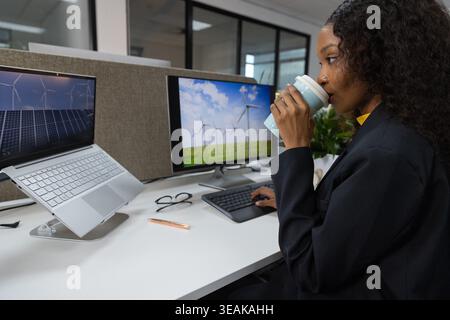 African American woman wearing dark blazer sipping mug and typing on keyboard at desk, copy space. Solar, wind, office, modern, laptop, monitor, glass Stock Photo