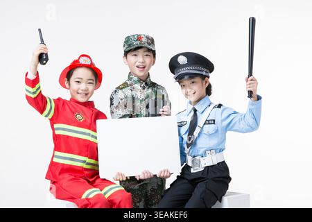 Three children in different costumes sit together and hold a blank sign, smiling at the camera. Stock Photo